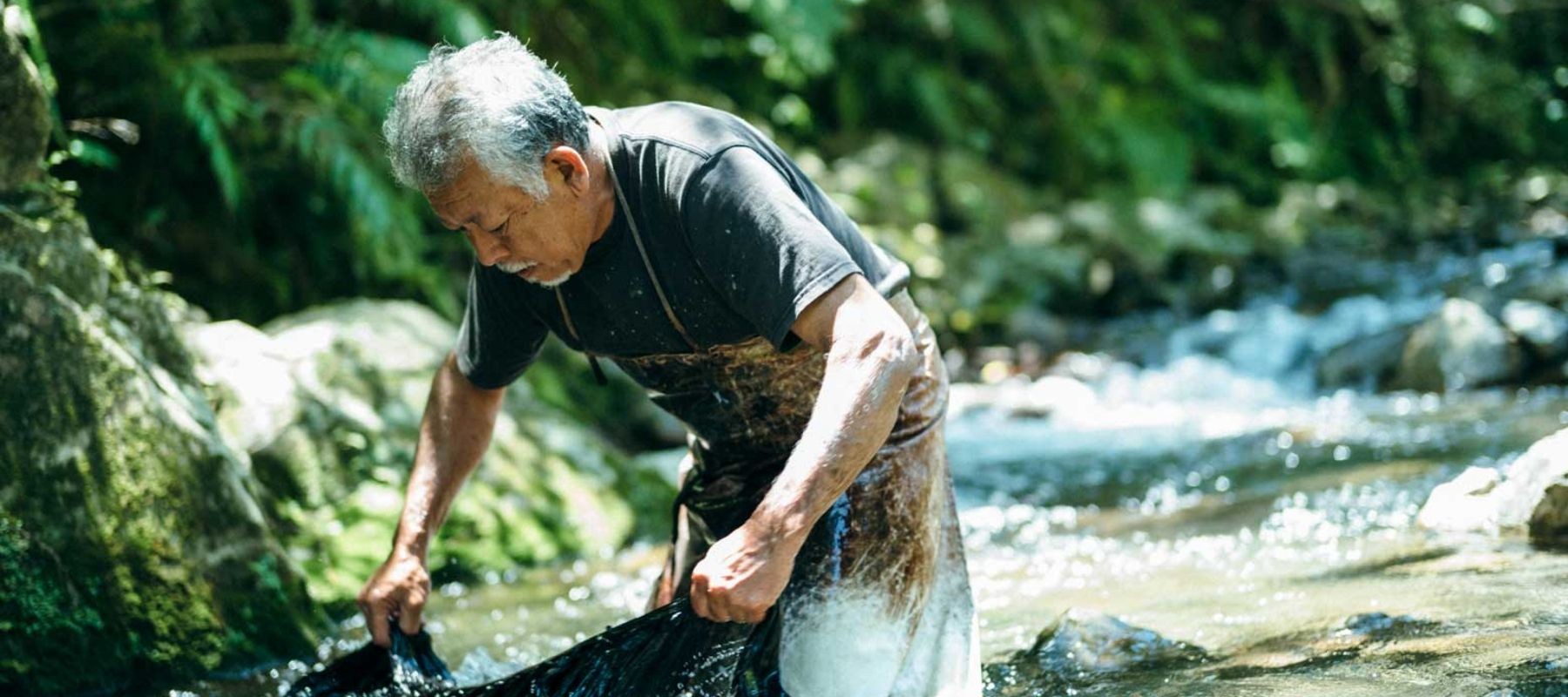 Person washing clothes in a river, surrounded by green vegetation and rocks.